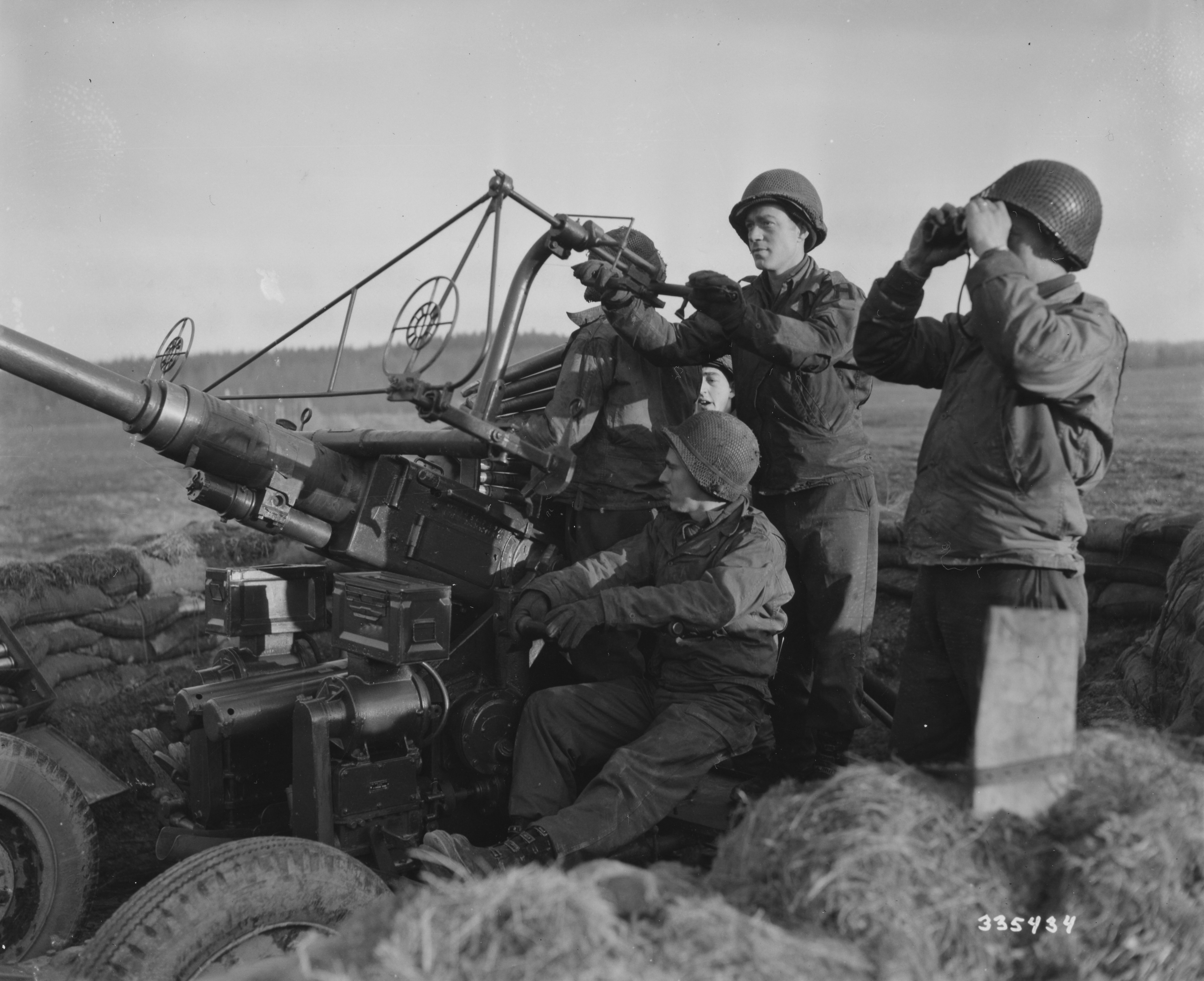 A U.S. First Army 40mm anti-aircraft gun crew alerted in a sandbagged pit near Eupen, Belgium, one man at the sight and another scanning the sky with binoculars, winter 1944 to 1945.